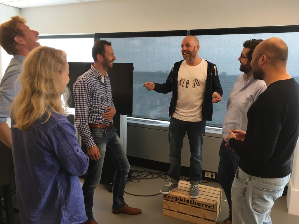 Zes mensen lachen om een spreker die op een houten kist met het opschrift Zeepkistborrel staat.